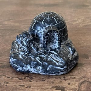 Op de afbeelding: Een kleine, zwart-witte sculptuur van een iglo met een figuur ervoor. De iglo heeft een koepelvormig dak met gesegmenteerde lijnen. De basis is getextureerd en de sculptuur staat op een houten oppervlak.