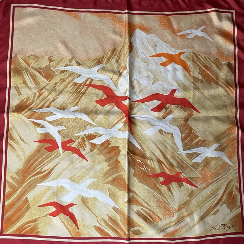 May include: Square silk scarf with a mountain landscape and flying birds in red, white, and orange. The scarf has a burgundy border and the text "La Plagne" in the bottom right corner.