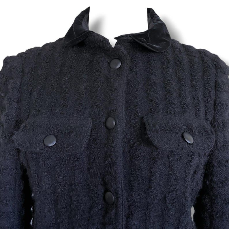 May include: A black textured jacket with a velvet collar and two front pockets. The jacket has four black buttons down the front and on the pockets. The fabric has a subtle vertical pattern.