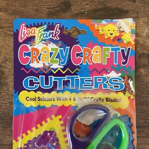 May include: A colourful package of Lisa Frank Crazy Crafty Cutters scissors. The package features the words "Crazy Crafty Cutters" in large, multi-coloured letters. The scissors have green handles and purple blades.
