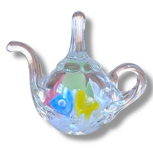 Puede incluir: Un objeto decorativo en forma de tetera de cristal transparente. El interior presenta diseños florales coloridos en rosa, azul, amarillo y verde. La tetera tiene un asa y un pico, y está diseñada como un artículo decorativo.