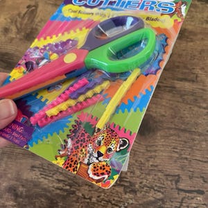 May include: A pack of colourful craft scissors with a green and purple handle and a pink handle. The package includes additional decorative blades in yellow, pink, and purple. The packaging features a jaguar illustration and the text "Cool Scissors With Blade."