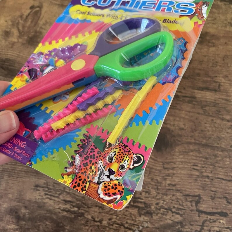 May include: A pack of colourful craft scissors with a green and purple handle and a pink handle. The package includes additional decorative blades in yellow, pink, and purple. The packaging features a jaguar illustration and the text "Cool Scissors With Blade."