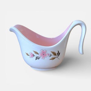 May include: White ceramic gravy boat with a pink interior and a curved handle. The exterior features a floral design with pink flowers and brown leaves. A vintage-style serving dish.