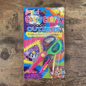 May include: A Lisa Frank Crazy Crafty Cutters set. The packaging is colourful with the text "Crazy Crafty Cutters" and "Cool Scissors With 4 Crafty Blades!". The scissors are pink, green, and purple.