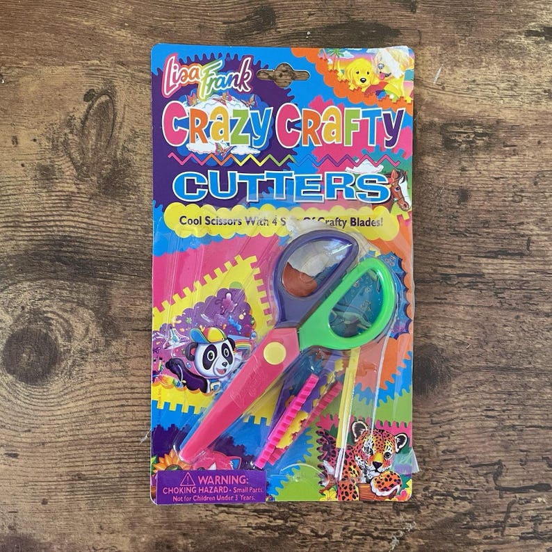 May include: A Lisa Frank Crazy Crafty Cutters set. The packaging is colourful with the text "Crazy Crafty Cutters" and "Cool Scissors With 4 Crafty Blades!". The scissors are pink, green, and purple.