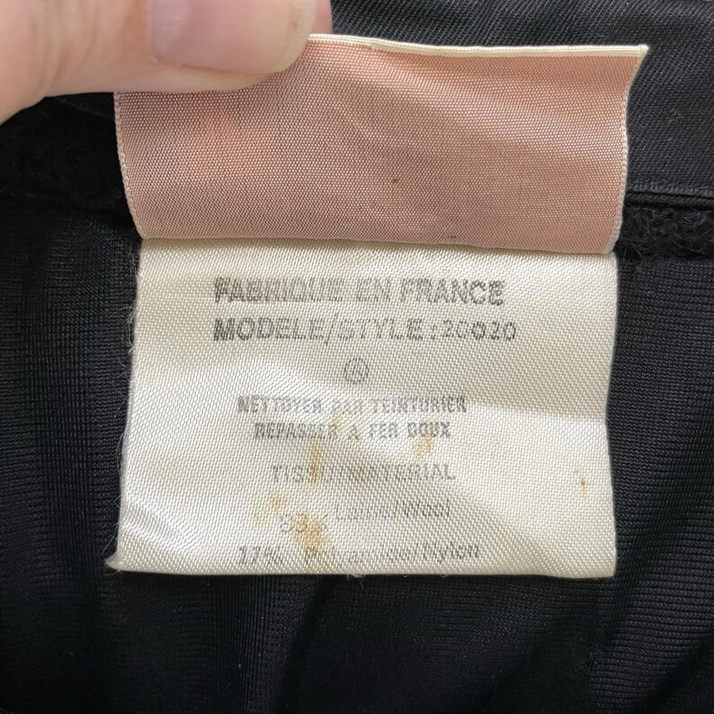 May include: Close-up of a clothing tag with text in French. The tag is white with black text and a pink top. The text includes "Fabriqu&eacute; en France" and "Mod&egrave;le/Style: 20020". The fabric is 83% wool and 17% polyamide nylon.