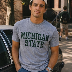 May include: A heather grey short-sleeved crew neck t-shirt featuring the words MICHIGAN STATE printed in dark green block lettering across the chest. The shirt is paired with blue denim jeans, a silver-tone wristwatch, and a white baseball cap.