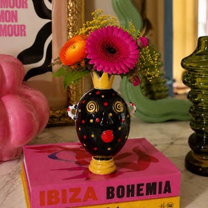 May include: A black ceramic vase with a yellow crown-shaped rim, yellow and red polka dots, and a stylized face design. The vase features two small handles and holds a bouquet of bright pink, orange, and yellow flowers, resting on two books.