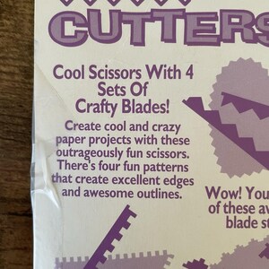 May include: A white package with purple text that reads "CUTTERS" at the top. The text below says "Cool Scissors With 4 Sets Of Crafty Blades!" and describes the scissors for paper projects.