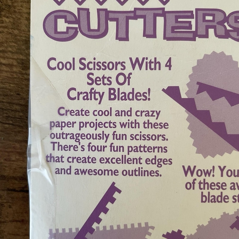 May include: A white package with purple text that reads "CUTTERS" at the top. The text below says "Cool Scissors With 4 Sets Of Crafty Blades!" and describes the scissors for paper projects.