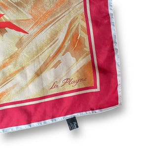 May include: A silk scarf with a red border and a cream-colored center featuring an abstract design with orange and red accents. The text "La Plagne" is printed in the center.