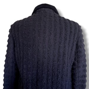 May include: A dark navy blue textured jacket with a black velvet collar. The jacket has a vertical ribbed pattern and is viewed from the back, showcasing the design and fabric texture. The garment appears to be a coat or blazer.