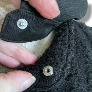 May include: Close-up of a black garment with a textured, fuzzy appearance. The garment features a black collar with a snap closure. The snap is silver and circular, with a decorative design around the center.