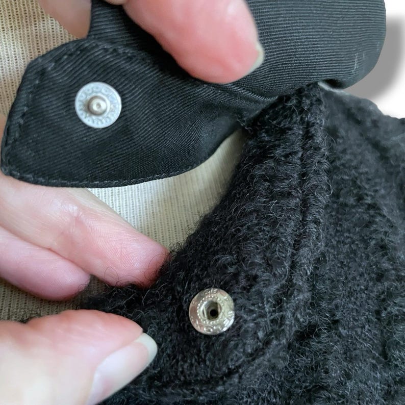 May include: Close-up of a black garment with a textured, fuzzy appearance. The garment features a black collar with a snap closure. The snap is silver and circular, with a decorative design around the center.