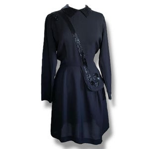 Peut inclure: Une robe noire vintage à manches longues et col. La robe présente une ceinture décorative à paillettes et des ornements sur l'épaule et la hanche. La jupe a un design superposé.