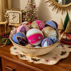 Puede incluir: Una colección de adornos navideños redondos en un cuenco de cerámica. Los adornos presentan texturas de terciopelo y purpurina en tonos rosa, morado, azul y dorado, decorados con abalorios, lentejuelas y pequeños brillantes en varios patrones.