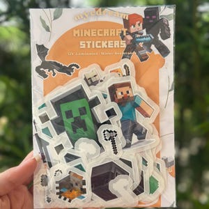 May include: A clear plastic package of Minecraft stickers. The package has the words "Minecraft Stickers" in orange and white text. The stickers feature various Minecraft characters and objects, including Steve, a Creeper, and an Enderman.