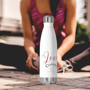 May include: White insulated water bottle with a silver cap and base. The bottle has the words "Love covers" in red and black script with a heart symbol. Designed to keep drinks cold for hours, it is a stylish hydration accessory.