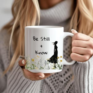 Op de afbeelding: Een witte keramische mok met een zwart silhouet van een vrouw die door een bloemenveld loopt. Op de mok staat de tekst "Be Still + Know".
