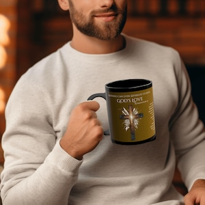 May include: Black coffee mug with the text "GOD'S LOVE" and a cross design. The mug also features the quote "NOTHING CAN EVER SEPARATE US FROM GOD'S LOVE ROMANS 8:39". The mug is held by a person.