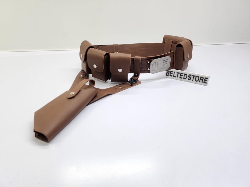 May include: A brown leather utility belt with multiple pouches and a holster. The belt has a silver buckle and is designed to be worn around the waist. The word "BELTEDSTORE" is printed on a white sign.
