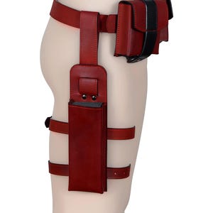 Peut inclure: Une ceinture en cuir rouge avec deux pochettes et un holster de cuisse. La ceinture a une bande noire et les pochettes ont des accents noirs.