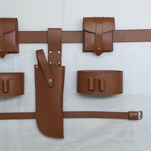 Puede incluir: Un cintur&oacute;n de cuero marr&oacute;n con dos bolsas de cuero marr&oacute;n, una funda de cuero marr&oacute;n y dos brazaletes de cuero marr&oacute;n. El cintur&oacute;n tiene una hebilla plateada y las bolsas tienen broches plateados.