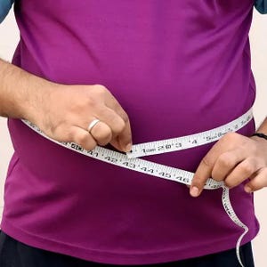 May include: A person wearing a purple shirt is using a white measuring tape to measure their waist. The tape is marked with both inches and centimeters, showing a measurement around 43 inches.