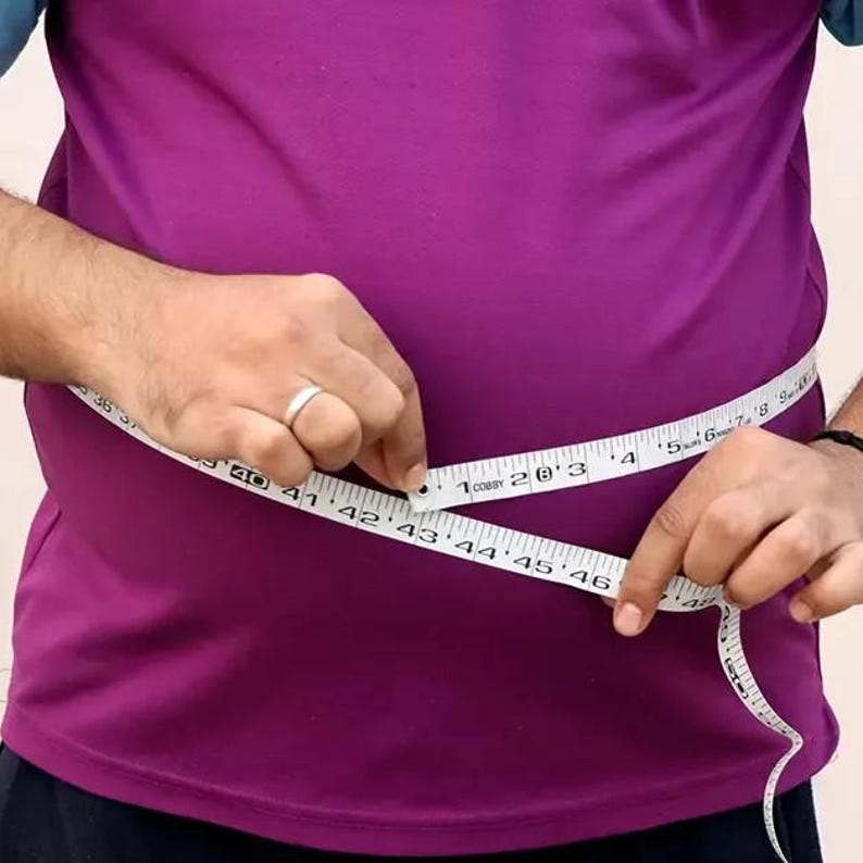 May include: A person wearing a purple shirt is using a white measuring tape to measure their waist. The tape is marked with both inches and centimeters, showing a measurement around 43 inches.