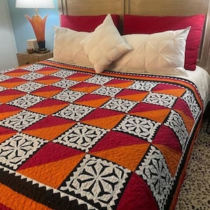 May include: A bed with a colorful quilt featuring a geometric pattern of red, orange, and white squares. The quilt is on a bed with white and red pillows. A wooden headboard and a lamp are visible in the background.