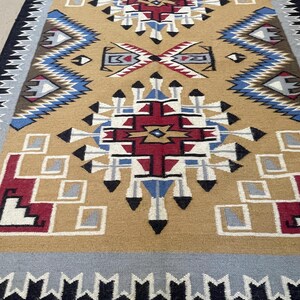 May include: A tan, geometric patterned rug with a central design in red, white, blue, and black. The rug features a border of gray, black, and white geometric shapes. The rug has a fringe along the bottom edge.