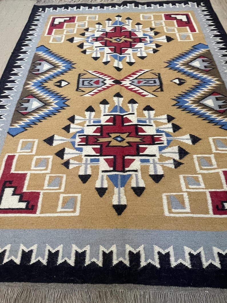 May include: A tan, geometric patterned rug with a central design in red, white, blue, and black. The rug features a border of gray, black, and white geometric shapes. The rug has a fringe along the bottom edge.