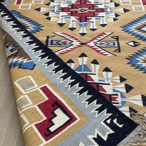 May include: A tan, geometric patterned rug with a folded edge. The rug features a central design with red, white, and blue accents. The border is black with white zigzags, and the edge is gray. The rug has a fringe.