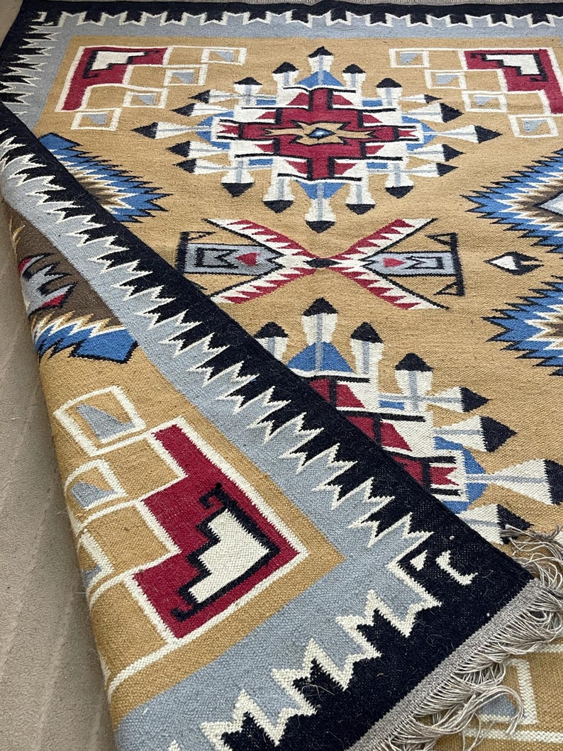 May include: A tan, geometric patterned rug with a folded edge. The rug features a central design with red, white, and blue accents. The border is black with white zigzags, and the edge is gray. The rug has a fringe.