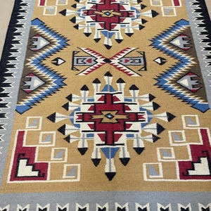 May include: A rectangular rug with a tan central field featuring geometric patterns in red, white, blue, and black. The rug has a black border with a gray and white patterned edge. The design includes diamond and square shapes.