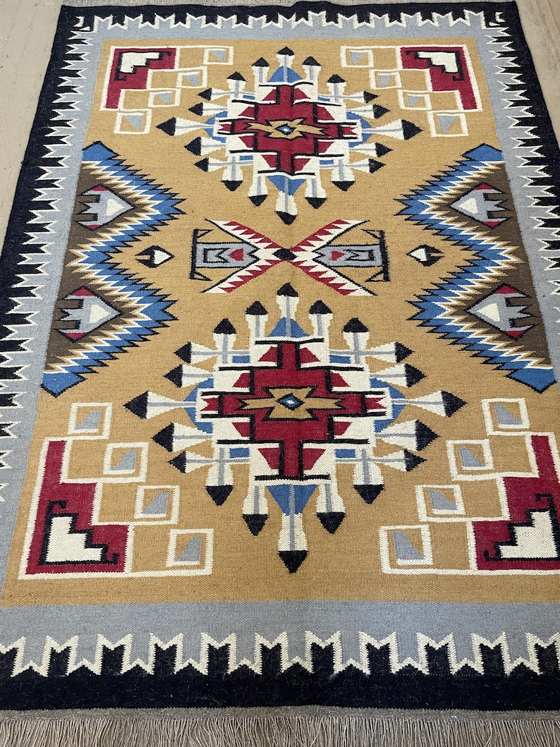 May include: A rectangular rug with a tan central field featuring geometric patterns in red, white, blue, and black. The rug has a black border with a gray and white patterned edge. The design includes diamond and square shapes.