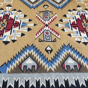 May include: A tan, geometric patterned rug with a central design featuring red, white, blue, and black accents. The rug has a border of black and gray triangles. The rug's design is symmetrical and features repeating patterns.