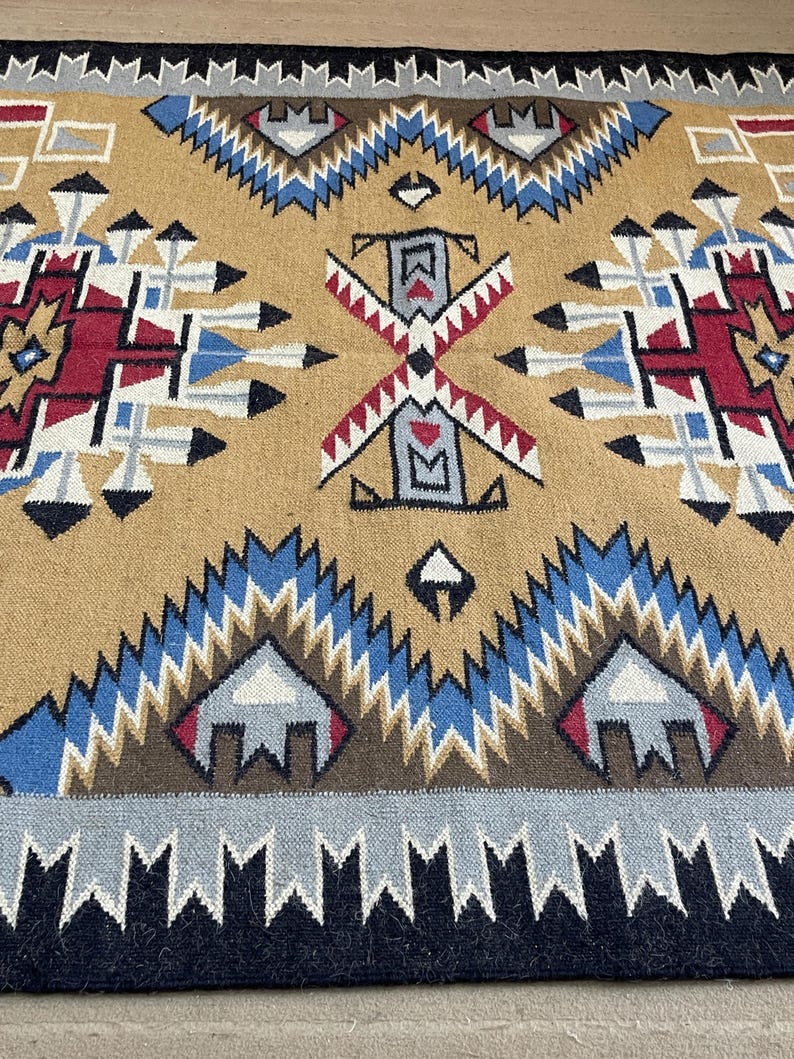 May include: A tan, geometric patterned rug with a central design featuring red, white, blue, and black accents. The rug has a border of black and gray triangles. The rug's design is symmetrical and features repeating patterns.