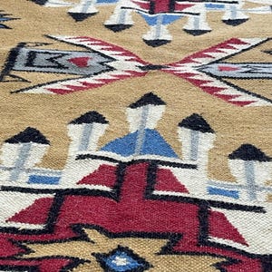 May include: Close-up of a woven rug with a geometric pattern. The rug features a tan background with a repeating design in red, black, blue, and white. The pattern includes diamond and arrow shapes.