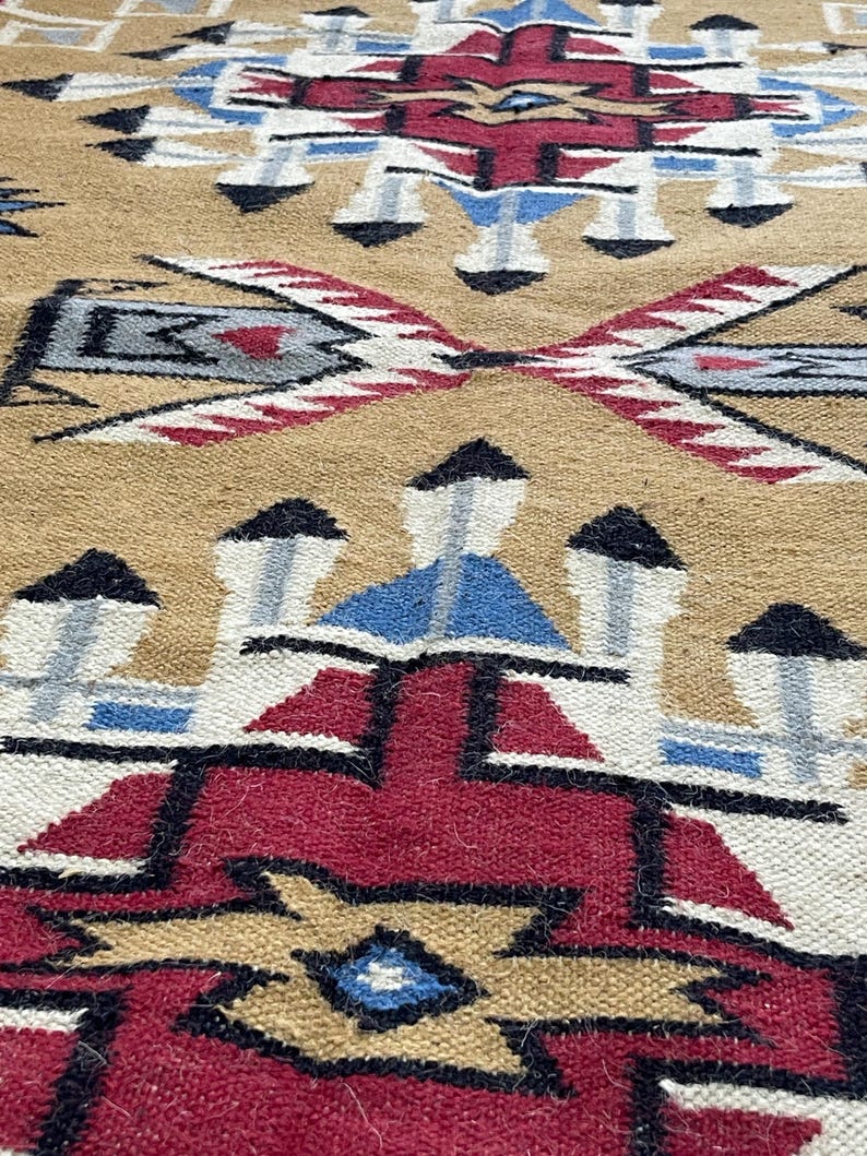 May include: Close-up of a woven rug with a geometric pattern. The rug features a tan background with a repeating design in red, black, blue, and white. The pattern includes diamond and arrow shapes.