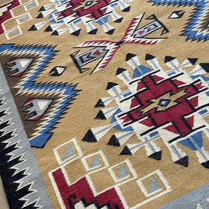 May include: A tan-colored woven rug with geometric patterns in red, blue, black, and white. The rug features repeating diamond and arrow designs, with a border of black and gray zigzags. The rug is a decorative floor covering.