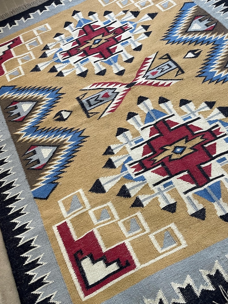 May include: A tan-colored woven rug with geometric patterns in red, blue, black, and white. The rug features repeating diamond and arrow designs, with a border of black and gray zigzags. The rug is a decorative floor covering.