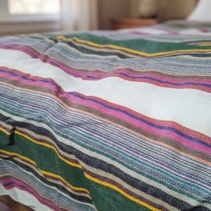 May include: A colorful woven blanket with horizontal stripes in shades of green, white, pink, purple, and yellow. The blanket is draped over a bed, showcasing its textured surface and vibrant design. The blanket is made of a soft material.