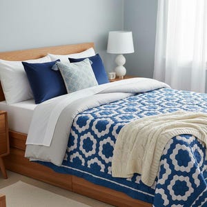 May include: A bedroom scene featuring a wooden bed frame with a blue and white patterned quilt. The quilt has a geometric design. Navy blue pillows and a white lamp complete the decor. A cream-colored throw blanket is draped over the quilt.