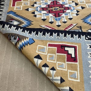 May include: A tan, blue, red, and white geometric patterned rug. The rug has a central design with a starburst pattern in red, white, and blue. The border is a repeating pattern of black and white triangles. The rug is folded over, revealing the underside.