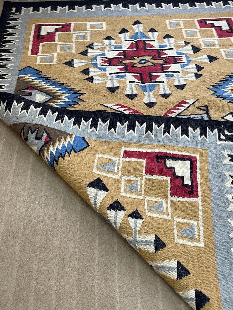 May include: A tan, blue, red, and white geometric patterned rug. The rug has a central design with a starburst pattern in red, white, and blue. The border is a repeating pattern of black and white triangles. The rug is folded over, revealing the underside.