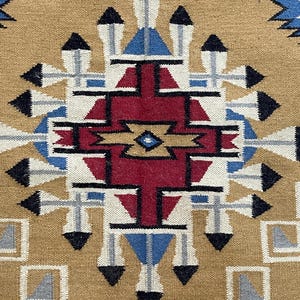 May include: Close-up of a woven textile with a geometric pattern. The design features a central red shape surrounded by blue, white, black, and tan elements. The background is a tan color, and the textile has a textured appearance.