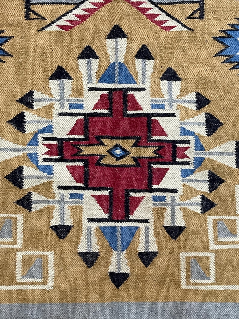 May include: Close-up of a woven textile with a geometric pattern. The design features a central red shape surrounded by blue, white, black, and tan elements. The background is a tan color, and the textile has a textured appearance.