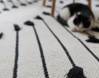 May include: A cream-colored rug with a repeating pattern of black lines and tassels. The rug is made of a woven material and has a textured surface. A black and white cat is resting on the rug.
